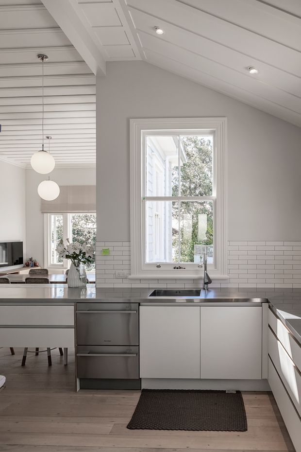Bright, refined kitchen with white flat-panel cabinetry, stainless-steel dishwasher, dark stone benchtops, and a white subway-tile splashback. A large framed window sits above the sink, while a central island extends toward an adjoining dining and living area. The room features white painted timber wall and ceiling panelling, pendant globe lights, pale timber flooring, and a pitched ceiling with recessed lighting.