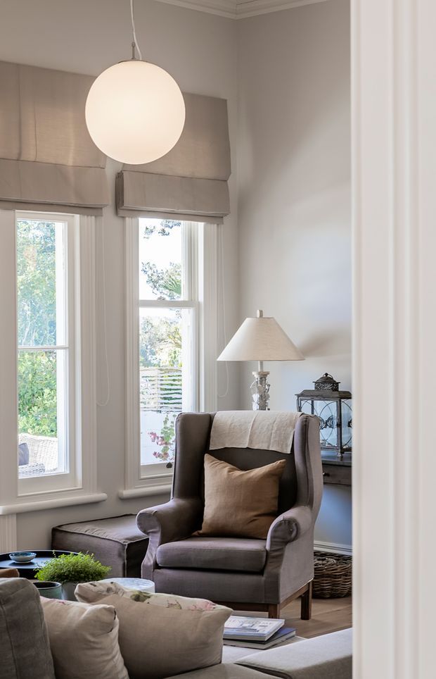 A refined, traditionally styled living room with tall sash windows, layered neutral Roman blinds, white-painted trim and walls, and warm timber flooring. The room features a large upholstered wingback armchair, neutral sofa seating, a side table with a lamp, a glowing spherical pendant light, and understated decorative accessories. The space feels calm, warm, and quietly elegant.