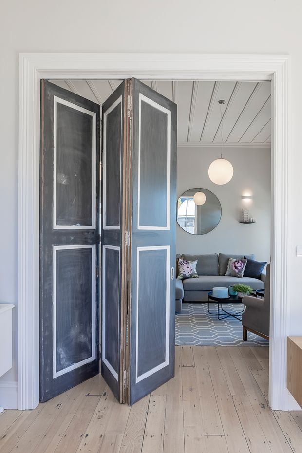 A bright interior view through open, dark-painted double doors with white inset panel mouldings. The foreground has wide pale timber floorboards and white decorative door and wall trim. Beyond is a refined living room with a grey sofa, patterned cushions, round black-framed mirror, pendant globe lights, a floating shelf, coffee table, armchair, and geometric grey rug. The ceiling features white painted boards with linear moulding details.