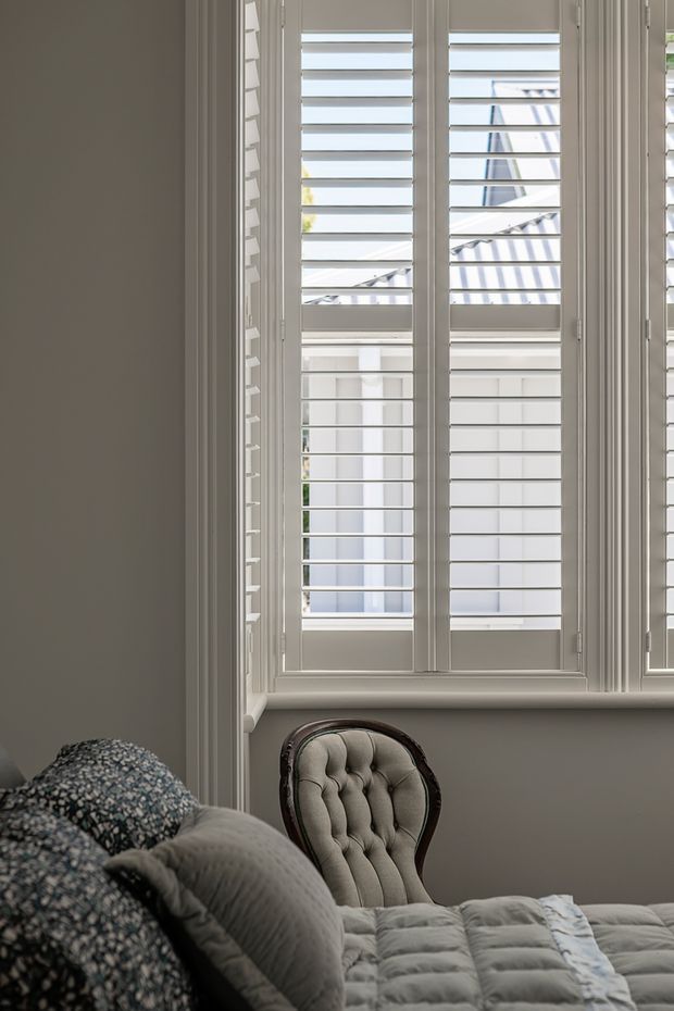 A softly lit bedroom interior featuring a large white-framed window with plantation shutters, grey walls and trim, and a partially visible upholstered bed with layered bedding and patterned cushions. A tufted upholstered accent chair sits beside the window. The room has a refined, traditional character with a muted neutral palette.