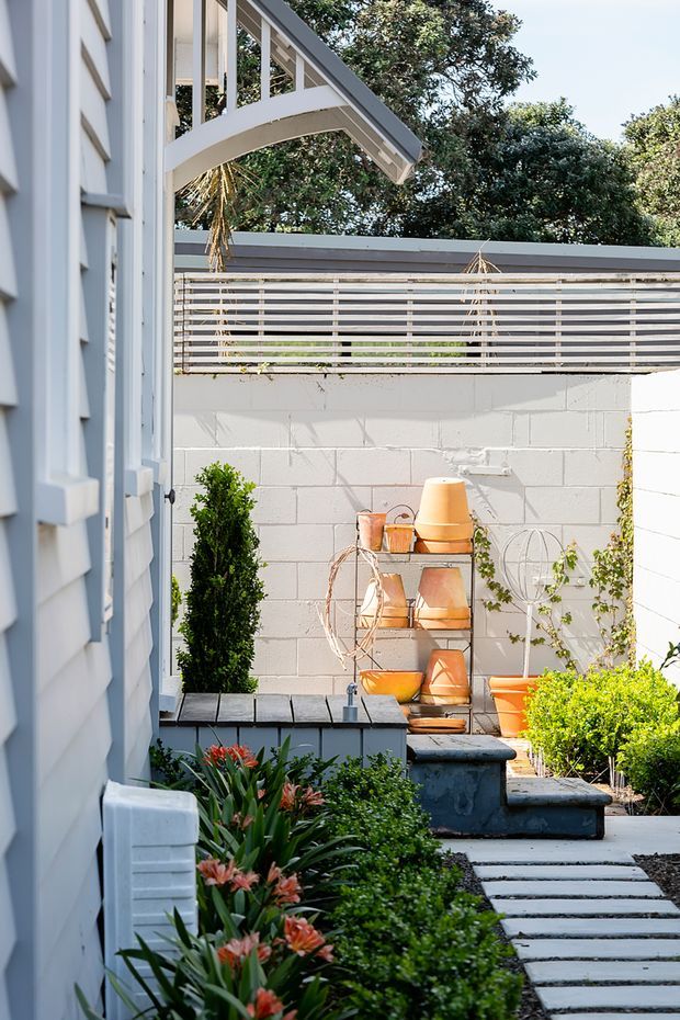 A bright, neatly landscaped courtyard or side garden beside a light-painted weatherboard building. The compact outdoor space features a white-painted concrete block boundary wall topped with horizontal slatted screening, a paved stepping-stone path, clipped shrubs, flowering plants, a dark raised planter or water feature, and a sculptural outdoor shelving display with terracotta pots. A projecting upper-level balcony or roof structure is visible above.
