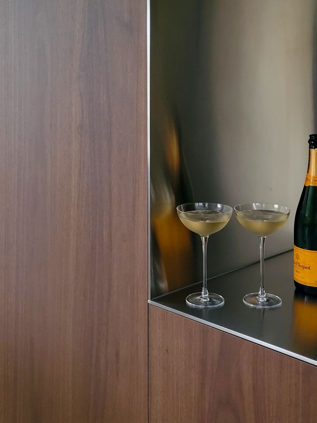 Close-up of a refined built-in drinks cabinet or bar niche, featuring dark timber veneer cabinetry, a mirrored or dark reflective rear panel, and a glossy dark benchtop. Two coupe glasses and a champagne bottle are displayed within the niche, creating an intimate, sophisticated presentation.