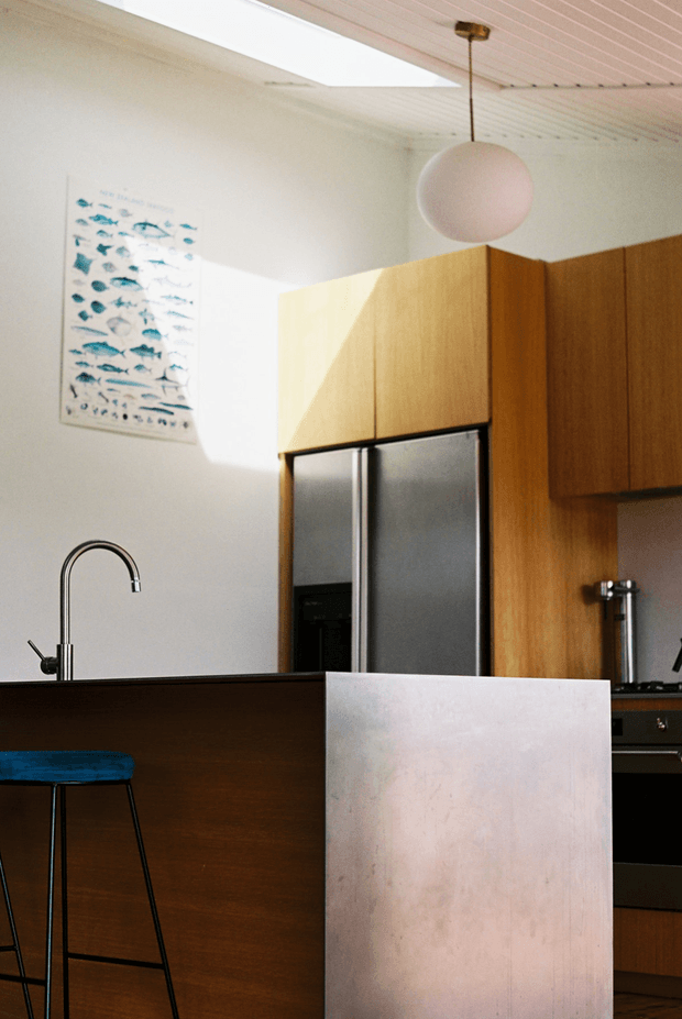 A warm, modern kitchen viewed across a dark timber island, with a tall curved chrome tap, integrated stainless-steel refrigerator, timber cabinetry, and a pale upholstered or panelled chair in the foreground. A blue upholstered bar stool sits at the island. The room has white walls, a white slatted ceiling, a large skylight, and a spherical pendant light. A fish illustration adds a small decorative accent.
