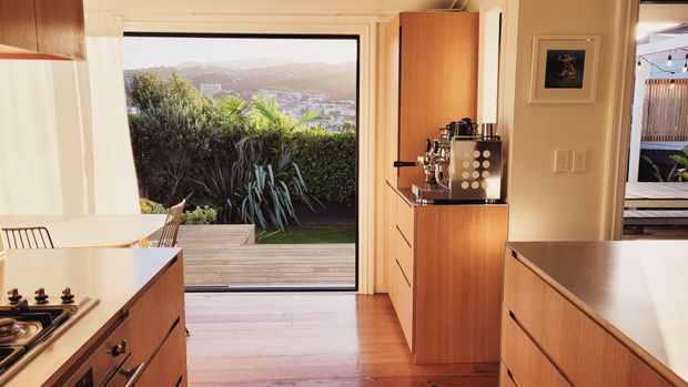 Warm modern kitchen with timber cabinetry, pale benchtops, integrated coffee station, and a large glazed opening leading to a timber deck and landscaped garden. A city and hillside view is visible beyond the planting. The space has clean-lined joinery, timber flooring, and strong indoor-outdoor connection.