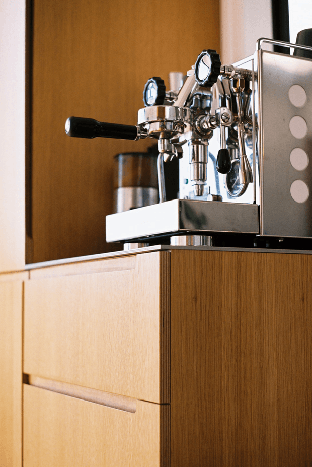 Close-up of a polished stainless-steel espresso machine with black portafilter and controls, positioned on a built-in timber cabinet or coffee station. The cabinetry has warm vertical wood grain, flat fronts, and recessed horizontal pulls. A timber wall panel forms the background, giving the image a warm, refined, minimal interior feel.