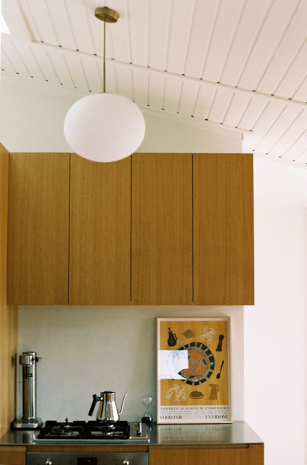A compact, warmly styled kitchen with flat-front walnut-toned upper cabinetry, a dark countertop, gas cooktop, stainless-steel appliances, and a framed artwork leaning against the splashback. The room has a white-painted timber-lined pitched ceiling and a spherical pendant light suspended from a brass-toned fitting. The composition emphasizes the cabinetry and ceiling details in a clean, mid-century-influenced interior.