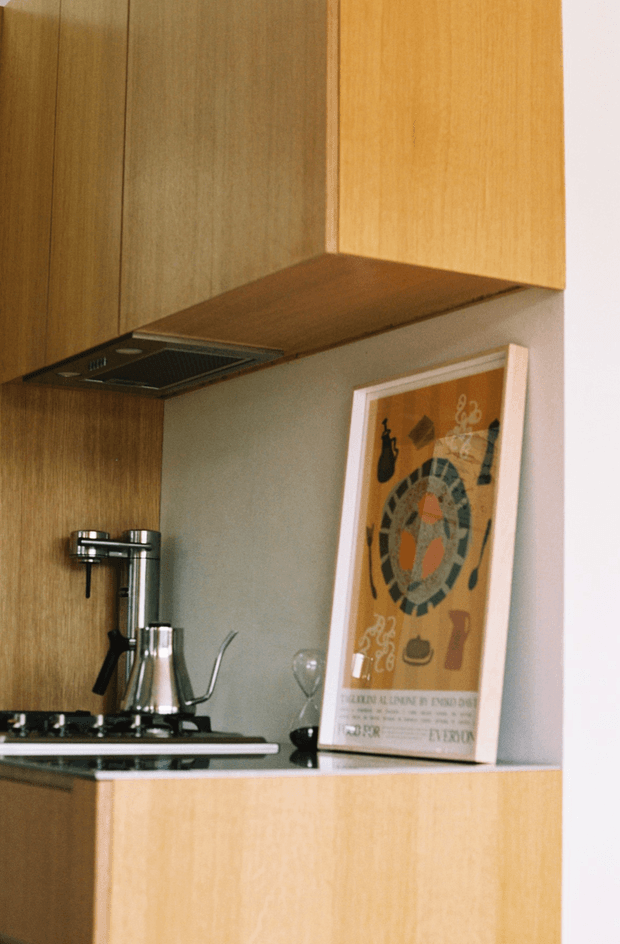 Close-up view of a compact modern kitchen or kitchenette with warm wood cabinetry, a dark induction cooktop and integrated rangehood, pale grey-green backsplash, and a light countertop. A chrome tap and coffee-making equipment sit on the benchtop, with framed artwork leaning against the backsplash.