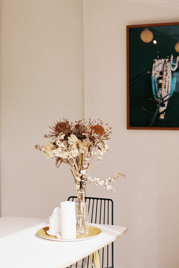A softly lit dining area with a white rectangular table, black metal spindle-back chairs, and a clear vase of dried floral arrangements. A round gold-toned tray holds white pillar candles on the table, while framed artwork with a dark background hangs on the warm off-white wall. The scene has a restrained, contemporary feel with warm natural light and muted earthy accents.