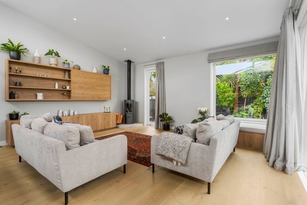 Bright, refined living room with two pale upholstered sofas arranged around a patterned rug and low coffee table. The room features light timber flooring, white walls and ceiling with recessed downlights, extensive windows with full-length curtains, and a wall-mounted timber storage and display unit. Indoor plants and a garden outlook add natural warmth.