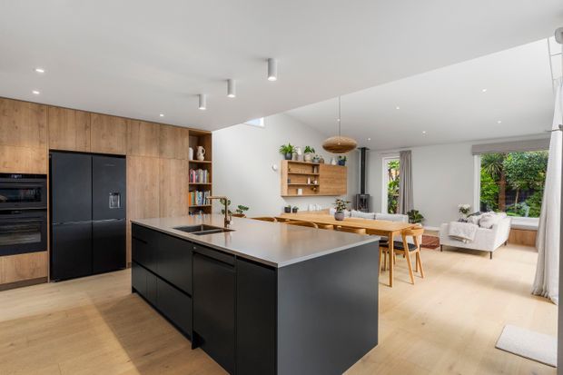 A bright open-plan kitchen and dining area with a large black island, integrated black appliances, and warm timber wall cabinetry. Light timber flooring continues into the adjacent living space, which features a dining table, pale upholstered seating, built-in timber shelving, pendant lighting, and garden views through large windows.