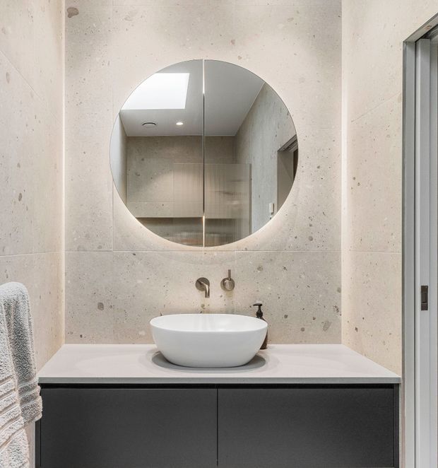 A contemporary bathroom vanity featuring a round wall mirror, white vessel basin, brushed metal wall-mounted tapware, pale concrete-look wall tiles, and dark flat-front cabinetry beneath a light benchtop. A skylight and recessed lighting brighten the compact space.