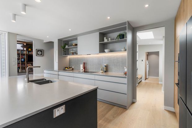 Bright contemporary kitchen featuring a large light-toned island with integrated double sink, dark island cabinetry, extensive grey built-in cabinetry, open shelving, and a softly illuminated textured backsplash. Pale timber flooring continues into the adjacent hallway, while a tall dark refrigerator is integrated into timber-toned cabinetry at right. A study or library area is visible through the opening at left.