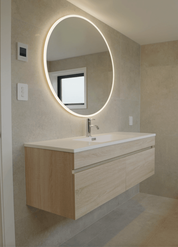 Contemporary bathroom vanity with a floating light-wood wall-mounted cabinet, white integrated benchtop and basin, chrome tapware, and a large round backlit mirror. The walls and floor are finished in warm beige-grey stone-look surfaces, creating a clean, refined, softly illuminated interior.
