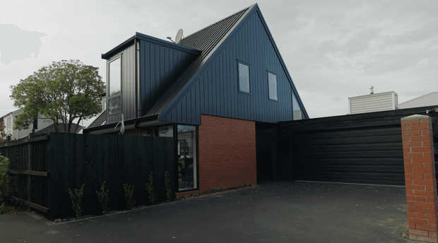 Exterior view of a compact contemporary dark-clad dwelling or extension with a steep gabled roof, vertical dark metal or timber-look cladding, exposed red brick walls, tall narrow windows, and a black garage door. A dark timber fence and asphalt driveway frame the foreground, with neighbouring buildings visible beyond.