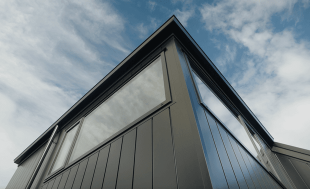 Low-angle exterior view of a contemporary building corner with dark vertical cladding, large reflective floor-to-ceiling glazing, and a sharply pitched or folded roofline against a cloudy sky.