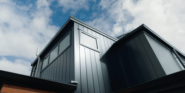 Low-angle exterior view of a contemporary multi-gabled building clad in dark vertical metal panels, with a small high-level window and visible roof junctions against a partly cloudy sky.