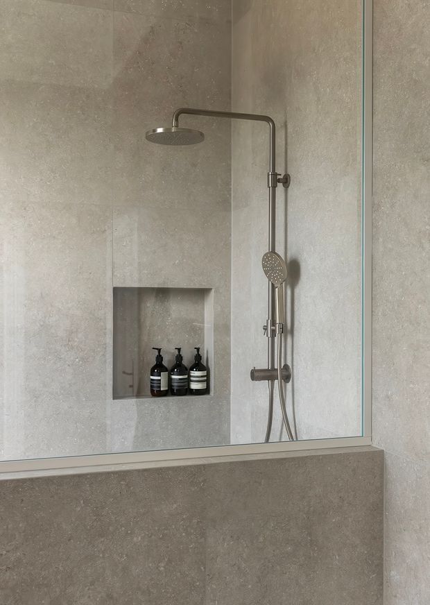 A minimalist walk-in shower enclosed by clear glass, with large grey-beige stone-look wall tiles, a recessed niche holding black bottles, and brushed metal rain and handheld shower fittings. The image has a calm, monochromatic, contemporary bathroom aesthetic.