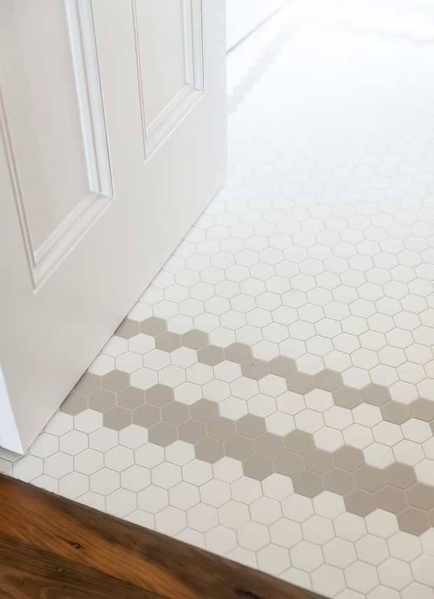 Close-up view of a bright interior threshold with a white raised-panel door opening onto a white hexagonal mosaic tile floor. The tiled surface includes subtle taupe and beige geometric bands, bordered by a dark stained timber floor at the bottom edge.