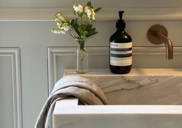 Close-up of a refined bathroom vanity area featuring a white marble or stone benchtop with subtle veining, a wall-mounted brushed bronze tap, decorative wall panelling, a glass vase with flowers, and a dark soap dispenser. A folded neutral towel drapes over the edge of a white freestanding or inset bathtub in the foreground.