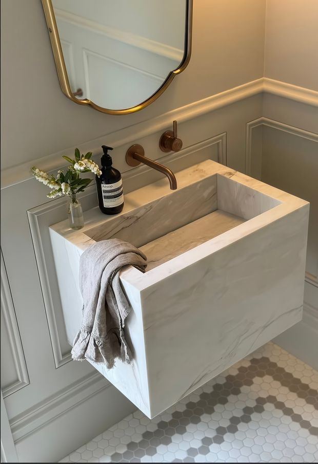 A refined bathroom vanity featuring a floating rectangular natural-stone basin with an integrated benchtop, wall-mounted brushed brass tapware, and a rounded brass-framed mirror. The walls are finished with grey-green painted decorative panel mouldings, while the floor uses small hexagonal tiles in light grey and beige tones. A folded linen towel, soap dispenser, and small floral arrangement add warmth.