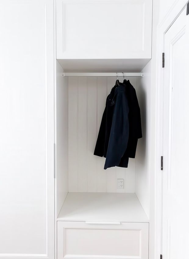 A compact built-in coat storage nook with white paneled cabinetry, a hanging rail with dark coats, vertical wall panelling, and a built-in bench or drawer unit below. The space is bright, clean, and highly integrated into the surrounding wall joinery.