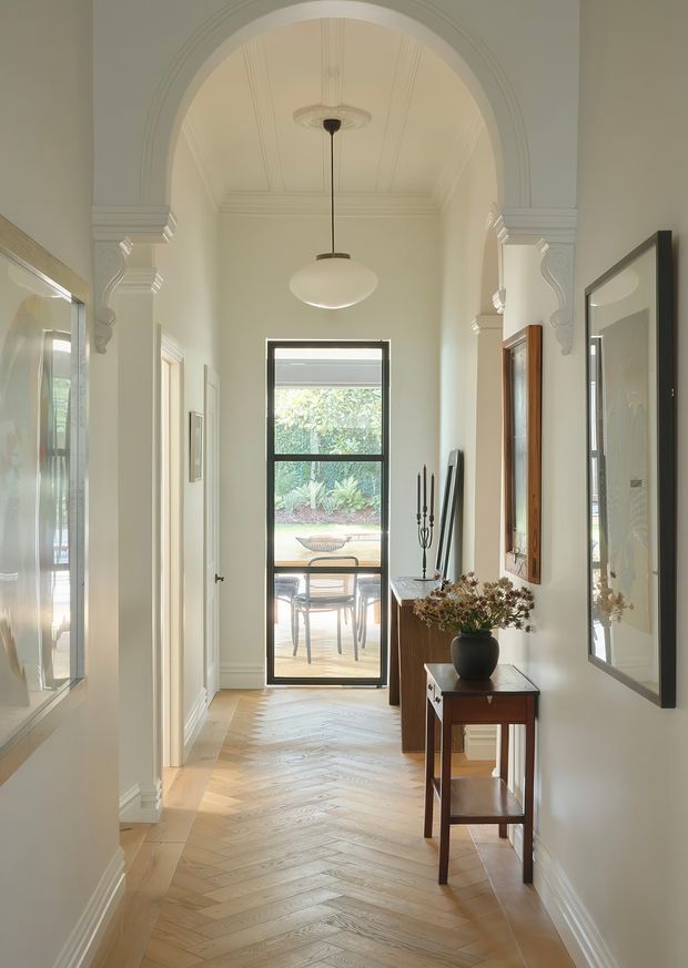 Elegant residential hallway with a long herringbone timber floor, pale walls, ornate arched opening, decorative plaster columns, coffered or panelled ceiling, and a central pendant light. A glazed door at the end opens to an outdoor seating area, while framed artwork, mirrors, a narrow timber console, and decorative objects line the hall.