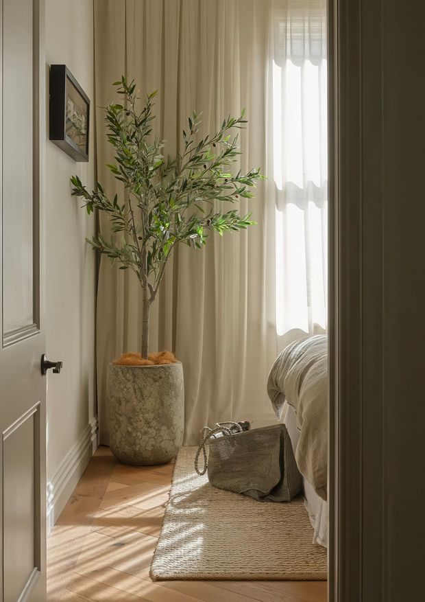 A warmly lit bedroom viewed through an open doorway, featuring a bed with neutral bedding, full-height beige curtains, a tall potted olive tree, timber flooring, a woven natural-fibre rug, and a textured stone-look planter. Decorative wall panelling and framed artwork add refined traditional detailing.