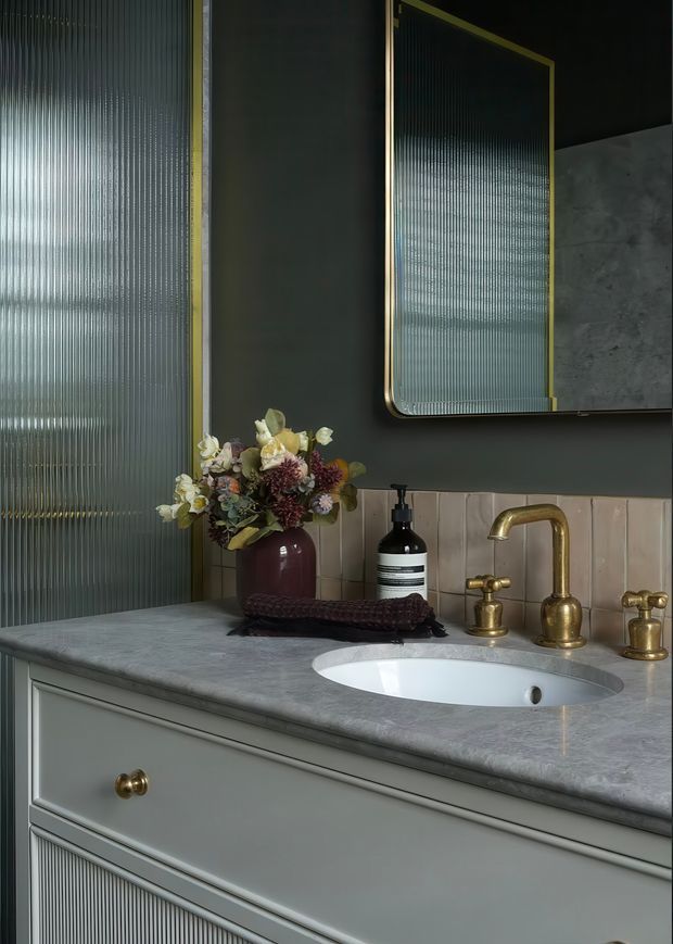 A moody, refined bathroom vanity with a grey painted cabinet, grey stone benchtop, inset white oval basin, and aged brass tapware. Ribbed translucent glass panels and a large ribbed-glass mirror add texture, while a pale tiled splashback and floral arrangement provide decorative detail.