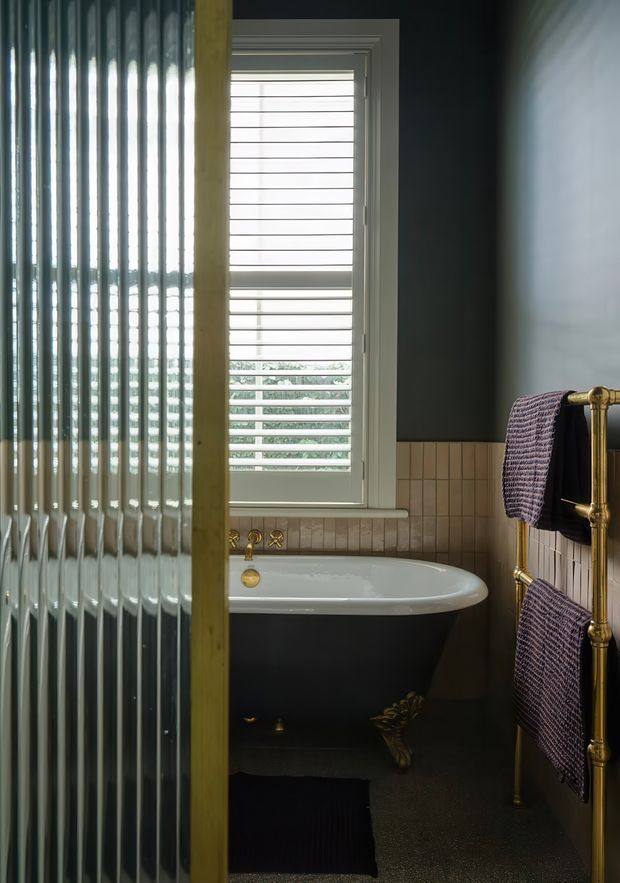 A compact, moody bathroom viewed through a ribbed glass partition. The room features a black freestanding clawfoot bathtub with white rim, brass plumbing and towel rail, pale square tiled wainscoting, dark painted upper walls, and a white shuttered window. Textured purple towels add a subdued accent.