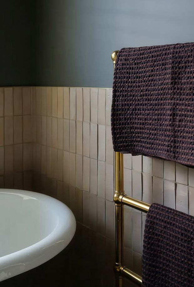 Close-up view of a refined bathroom corner featuring a curved white freestanding bathtub, pale rectangular wall tiles with dark grout, a dark painted upper wall, and a brass heated towel rail holding textured dark mauve towels. The lighting is subdued and atmospheric, creating a moody, boutique-hotel feel.