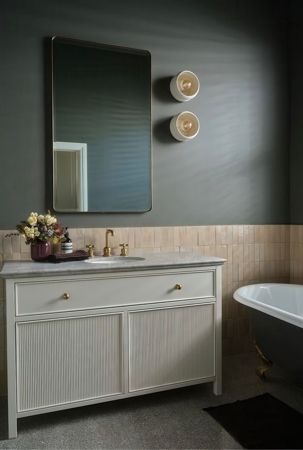 A moody, refined bathroom featuring a fluted pale vanity with brass knobs, a grey stone benchtop, twin basin taps, a large rounded-rectangle mirror, and warm beige vertical wall tiles. A freestanding bathtub with brass feet is partially visible at right, with soft wall lighting and floral styling adding a classic, elegant feel.