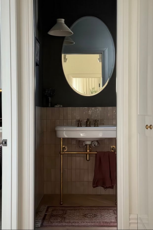 A compact, elegantly styled powder room viewed through an open doorway. The space features a wall-mounted white ceramic basin with exposed brass plumbing, dusty mauve-brown rectangular wall tiles, a matte black upper wall, a large arched mirror, and a pleated pendant light. A patterned rug and burgundy hand towel add warmth and character. White paneled door trim frames the room.