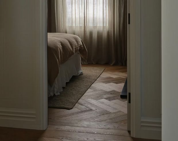 A softly lit bedroom viewed through an open doorway. The room features a bed with a taupe or beige rumpled coverlet, sheer full-height curtains, a muted textured area rug, and a distinctive light timber herringbone floor. White painted walls, doorway trim, and skirting create a calm, understated interior.