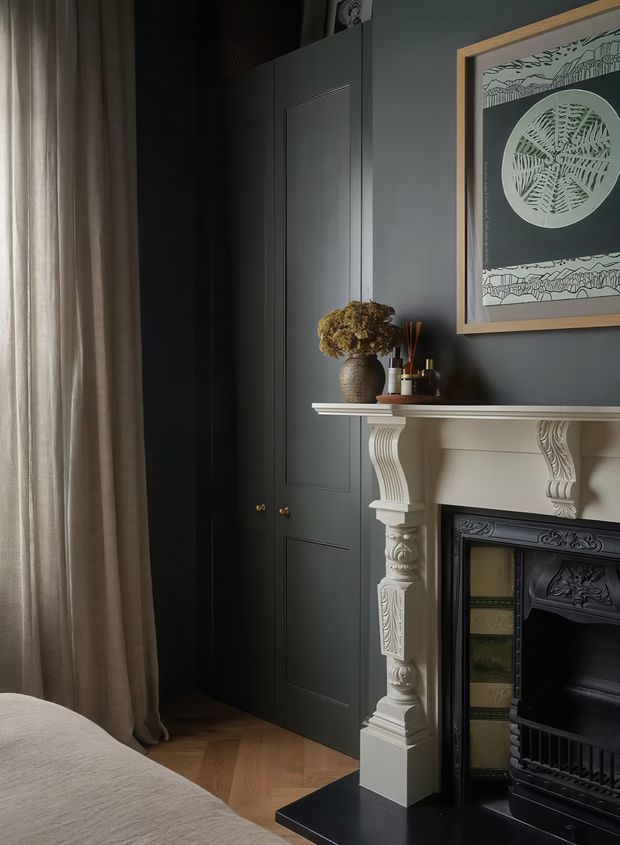 A moody, refined bedroom corner featuring dark blue-grey panelled built-in cabinetry, a decorative white carved mantel and ornate black fireplace surround. A bed with neutral bedding is partially visible in the foreground beside full-height taupe curtains. Framed botanical artwork, a ceramic vase with dried foliage and small decorative objects sit on the mantel. Herringbone timber flooring adds warmth.