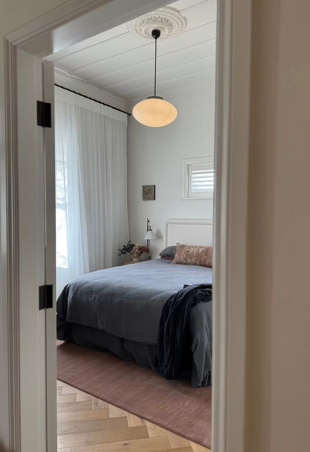 A serene bedroom viewed through an open doorway, featuring a neatly made bed with layered grey-blue bedding, a soft pink-beige rug, sheer floor-length curtains, a small shuttered window, and a warm globe pendant hanging from an ornate ceiling rose. The room has pale walls, decorative ceiling panelling, classic trim, and timber herringbone flooring extending into the foreground.