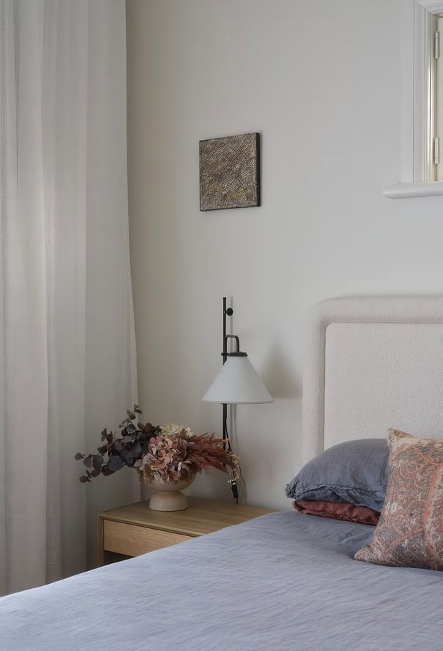A softly lit, calming bedroom corner featuring a partially visible upholstered bed with a light textured headboard, blue-grey bedding, and patterned cushions. A timber bedside table holds a ceramic vase with dried flowers and a simple white-shaded black wall-mounted lamp. Pale neutral walls, sheer curtains, and a small abstract artwork create a restrained, serene interior.