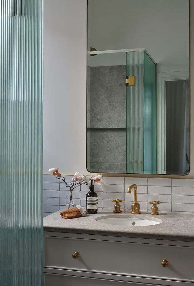 A refined bathroom vanity with a grey stone benchtop, inset white basin, brushed brass tapware, grey painted cabinetry, and white subway-tile splashback. A large rounded-rectangle mirror reflects a grey tiled shower enclosure with ribbed translucent glass and brass hardware. Ribbed glass is also visible in the foreground, adding texture and privacy. The palette is soft grey, white, muted green, and warm brass with a classic yet contemporary feel.