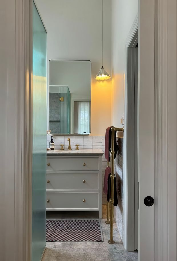 A compact, refined bathroom viewed through a partially open doorway. The room features a pale grey vanity with brass knobs and fittings, a light stone benchtop, white horizontal wall tiles, a large rounded-rectangle mirror, a decorative warm pendant light, and a freestanding brass towel rail with burgundy towels. A patterned floor mat sits over a stone-look floor, while a green-tinted fluted glass door or screen is visible at the left.