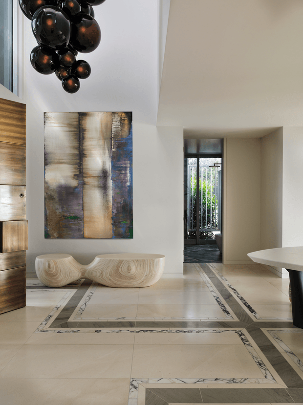 A refined contemporary entry hall with a pale stone-tile floor arranged in geometric rectangular inlays with dark veining. White walls and a high ceiling frame a dramatic cluster of glossy black pendant spheres, a large abstract artwork, and a sculptural light-wood console. A timber pivot door is partially visible at left, while a narrow glazed opening leads toward a garden or courtyard. The space has a luxurious, gallery-like appearance with restrained neutral finishes and bold sculptural accents.