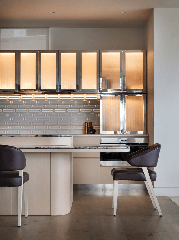 A sophisticated contemporary kitchen with a compact peninsula or island, integrated cabinetry, and a distinctive illuminated textured tile backsplash. The upper cabinets feature translucent ribbed glass fronts with warm internal lighting and dark metal frames. Pale wood-toned lower cabinetry, a light benchtop, dark leather dining chairs, and a timber floor create a warm, refined atmosphere.
