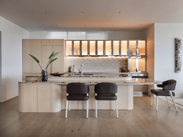 Contemporary open-plan kitchen dominated by a large curved island with integrated seating, pale timber cabinetry, a dark stone benchtop, and a reflective metallic tiled splashback. Full-height light cabinetry and illuminated upper display cabinets line the rear wall, while warm recessed lighting and timber flooring create a refined, inviting atmosphere. A small dining or breakfast seating area is visible at the island.