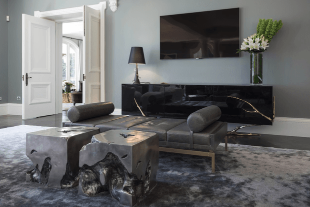 A refined living room with cool grey-blue walls, white architectural trim, dark glossy built-in cabinetry, and a wall-mounted television. A low upholstered sofa with cylindrical bolster cushions faces the TV, while two sculptural metallic coffee tables sit on a large grey patterned rug. A doorway opens to another bright interior space, and a tall vase with greenery provides a natural accent.