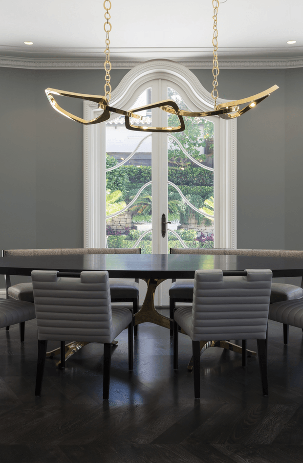 A refined dining room with a long dark dining table, upholstered grey dining chairs, and a sculptural gold pendant light suspended in front of an ornate arched glass door. The room features dark timber flooring, grey walls, detailed ceiling cornices, and traditional white architectural mouldings, creating a sophisticated blend of classic architecture and contemporary furnishings.
