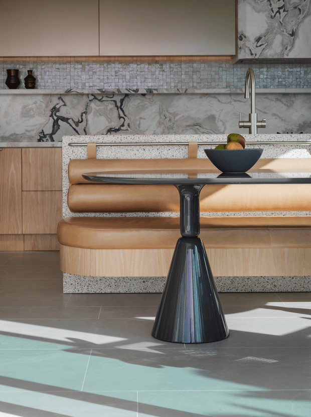 A refined contemporary kitchen viewed from the dining area, featuring light timber cabinetry, a large veined stone splashback, a small mosaic tile backsplash, and a terrazzo island benchtop. In the foreground is a sculptural black pedestal dining table with a bowl, while a curved tan upholstered bench wraps around the island. Strong sunlight casts geometric shadows across the tiled floor.