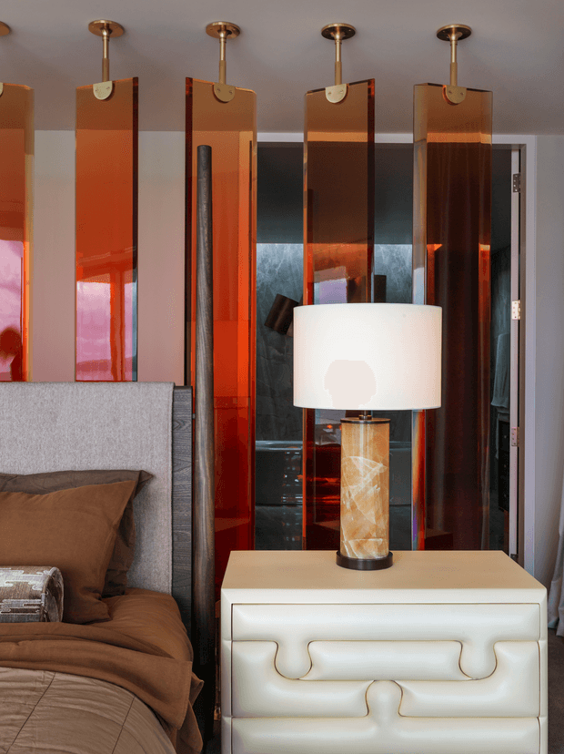 A close-up of a richly styled bedroom corner featuring a grey upholstered bed with layered brown bedding and pillows, a white sculptural bedside chest, and a table lamp with a translucent stone-like base and white drum shade. Behind the bed is a dramatic feature wall made from tall, glossy amber and red translucent panels suspended from the ceiling, with dark reflective surfaces and a doorway visible beyond. The palette is warm, luxurious, and highly contrasting, with reflective materials and bold geometric detailing.