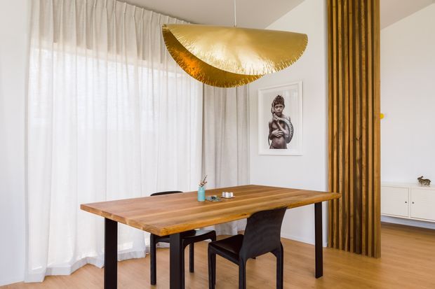 A refined dining area with a solid timber dining table and dark chairs, set against sheer full-height curtains and pale walls. A sculptural gold pendant light hangs above the table, while a vertical timber-slat screen separates the dining area from an adjacent living space with built-in white cabinetry. The room has warm timber flooring, restrained furnishings, and a framed artwork.