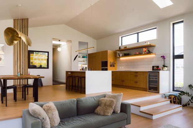 Bright open-plan living, dining and kitchen interior with warm timber flooring and cabinetry, a white island, tiled kitchen splashback, and large windows providing abundant natural light. The foreground contains a contemporary grey sofa, while the dining area, kitchen, bar seating and a small set of steps are visible. Vertical timber battens and a sculptural brass pendant add distinctive feature elements.