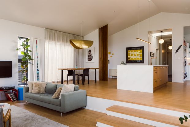 Bright open-plan living and dining area with a contemporary kitchen beyond a white island. The room features warm timber flooring, a pale grey sofa, sheer curtains, a sculptural gold pendant light, timber vertical screening, built-in cabinetry, and stepped changes in floor level connecting the living zone to the kitchen and entry.
