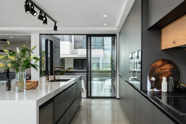 A contemporary open-plan kitchen with a large white stone island, dark integrated cabinetry, black-framed glazed doors, and polished concrete flooring. Track lighting and recessed ceiling lights illuminate the space, while timber shelving and decorative objects add warmth. The kitchen opens toward a bright courtyard or exterior view.