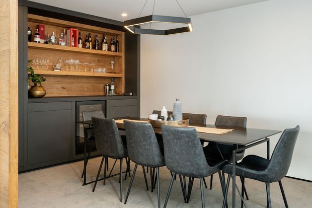 Contemporary dining and entertaining area featuring a long dark dining table surrounded by upholstered chairs, with a built-in dark bar cabinet and open timber shelving displaying bottles and glassware. A geometric pendant light hangs above the table, while light walls and flooring create a restrained, refined atmosphere.